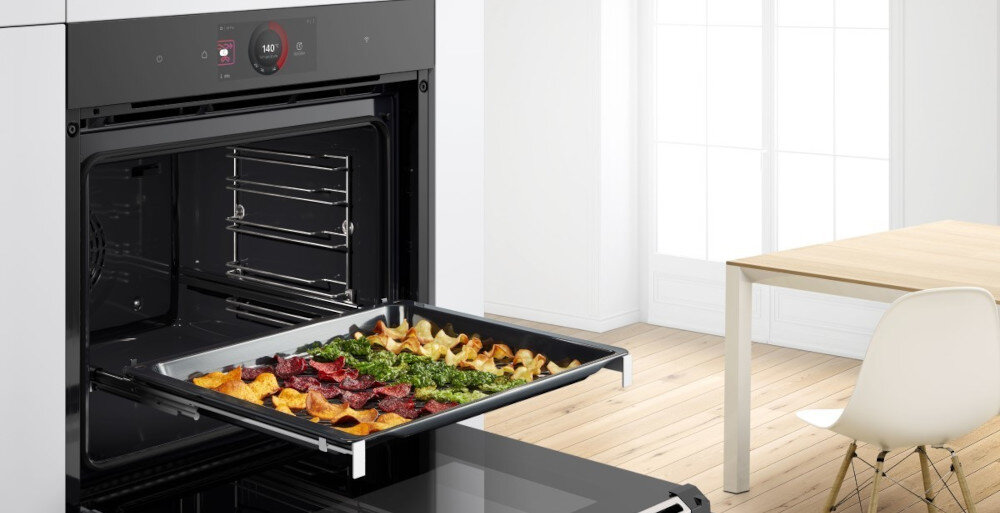 Otwarty czarny piekarnik z wysuniętą blachą, na której znajdują się różnorodne pieczone warzywa. W tle jasne wnętrze z drewnianą podłogą i stołem.