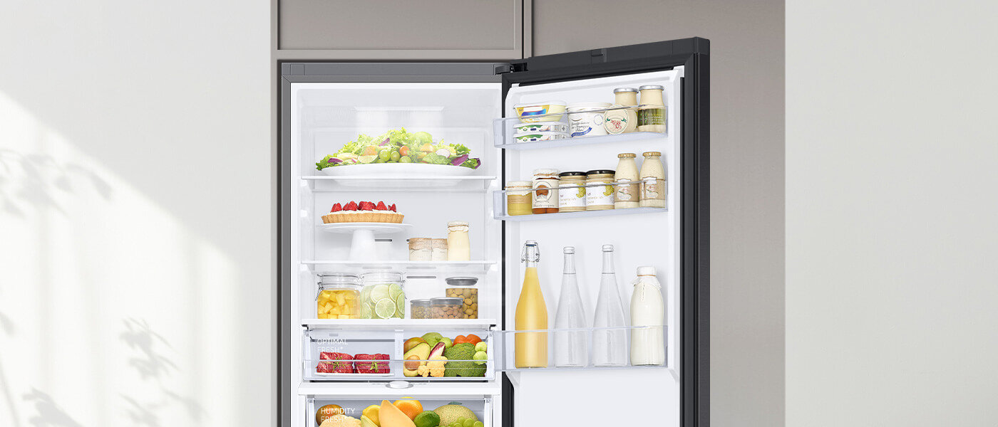 lodówka z dolnym zamrażalnikiem rb34c603cb1 posiada pojedynczy system chłodzenia All around cooling Otwarta lodówka z widocznymi półkami wypełnionymi różnorodnymi produktami spożywczymi, takimi jak warzywa, owoce, napoje i słoiki. Wnętrze jest jasno oświetlone, a drzwi lodówki mają dodatkowe półki na mniejsze produkty.