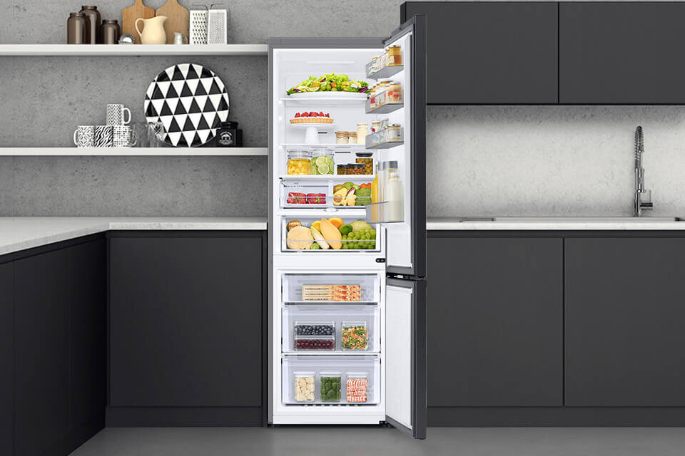Otwarta lodówka w kuchni, wypełniona różnorodnymi produktami spożywczymi, takimi jak owoce, warzywa i napoje. W tle widoczne są ciemne szafki kuchenne i półki z dekoracjami.