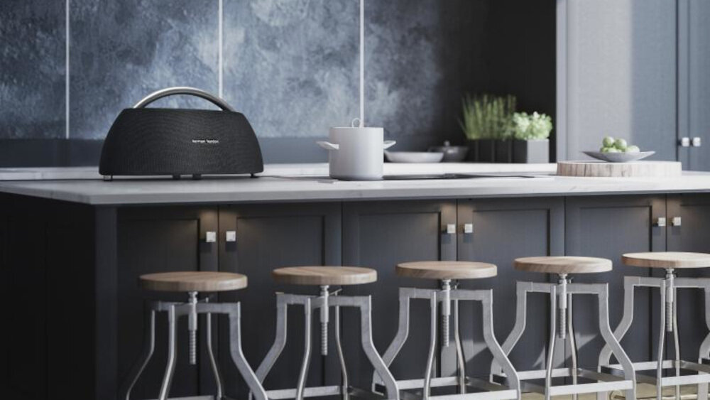 Na blacie kuchennym stoi czarny głośnik z logo \'harman/kardon\'. W tle widoczne są doniczki z roślinami i naczynia kuchenne.