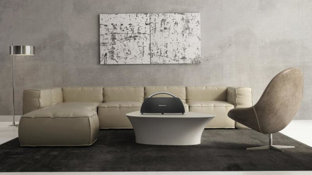 W nowoczesnym salonie na białym stoliku znajduje się czarny głośnik z logo \'HARMAN KARDON\'. W tle widoczna jest beżowa sofa, abstrakcyjny obraz na ścianie i brązowy fotel.