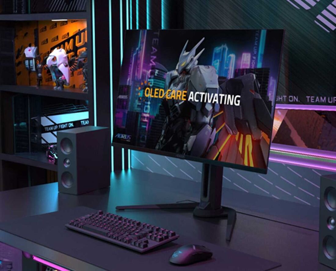 Na biurku znajduje się monitor z wyświetlonym obrazem robota i tekstem \'OLED CARE ACTIVATING\'. Obok monitora widoczne są głośniki, klawiatura i myszka, a w tle półki z figurkami.