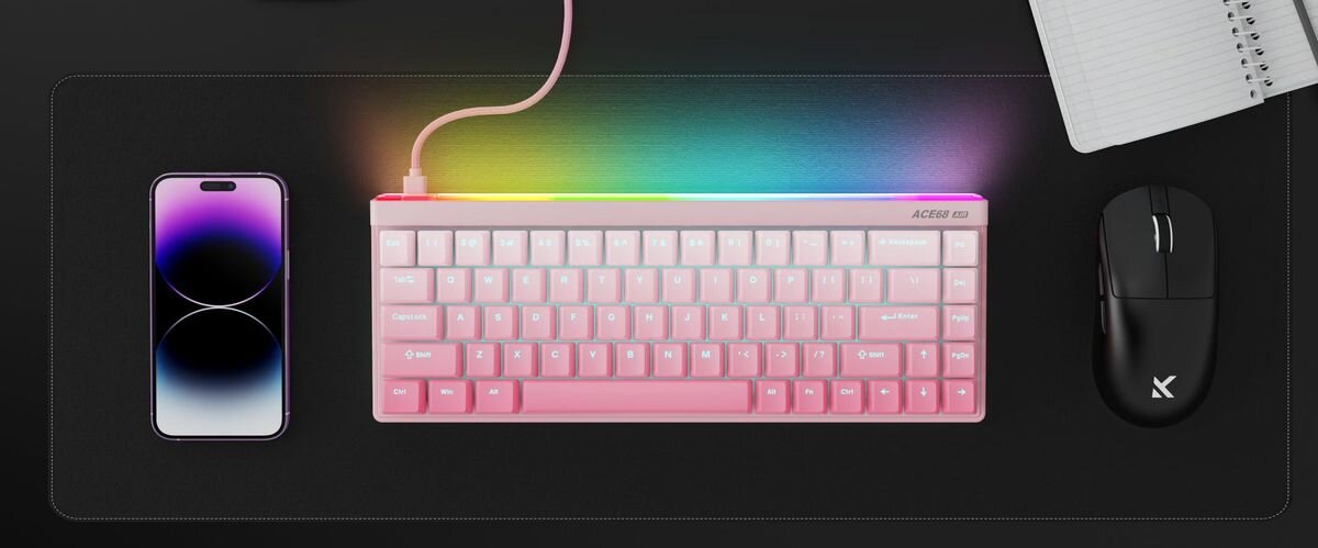 Na biurku znajduje się różowa klawiatura z podświetleniem RGB, obok niej leży czarny smartfon i myszka z logo \'X\'. W tle widoczny jest otwarty notatnik.