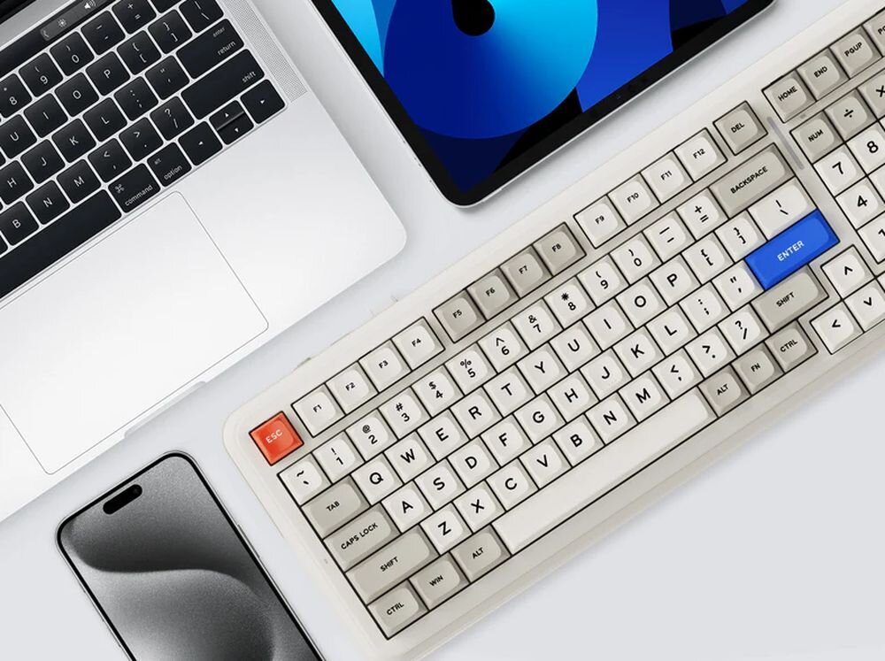 Na obrazie widoczna jest klawiatura o retro wyglądzie z białymi i szarymi klawiszami oraz niebieskim klawiszem ENTER. Obok klawiatury znajdują się laptop, tablet i smartfon.