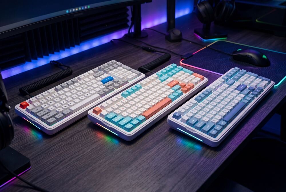 Na biurku znajdują się trzy klawiatury mechaniczne z różnokolorowymi klawiszami, każda z podświetleniem RGB. W tle widoczny jest monitor oraz akcesoria komputerowe.