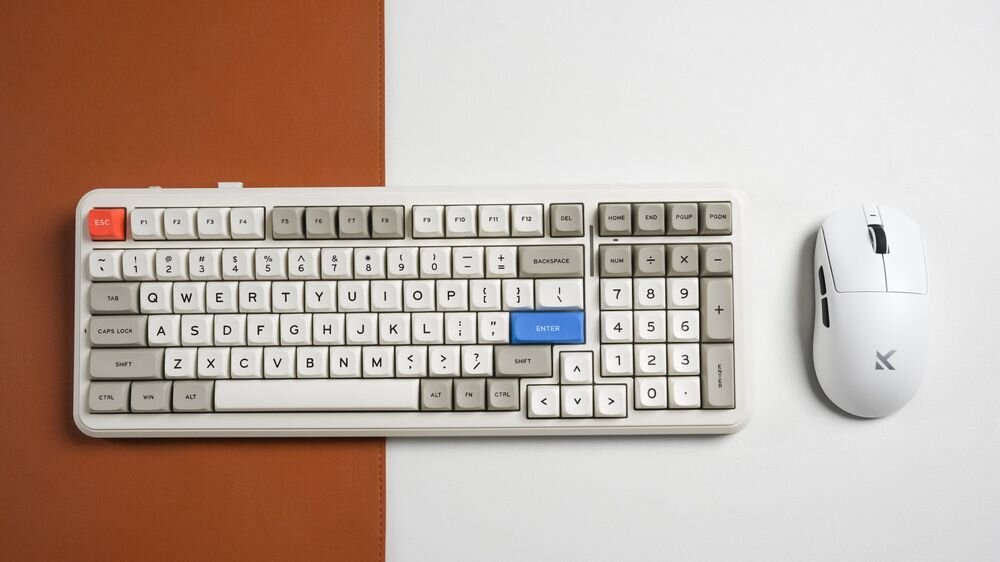 Na obrazie znajduje się biała klawiatura z kolorowymi klawiszami, w tym czerwonym klawiszem ESC i niebieskim klawiszem ENTER, oraz biała mysz z logo w kształcie litery X. Tło jest podzielone na brązową i białą część.