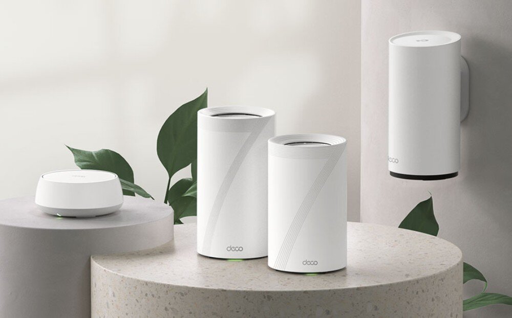Router TP-LINK Deco BE22 Cztery różne urządzenia sieciowe o kształcie walców i dysku ustawione na okrągłych podestach w otoczeniu roślin, rozbudowa, kompatybilność, skalowanie
