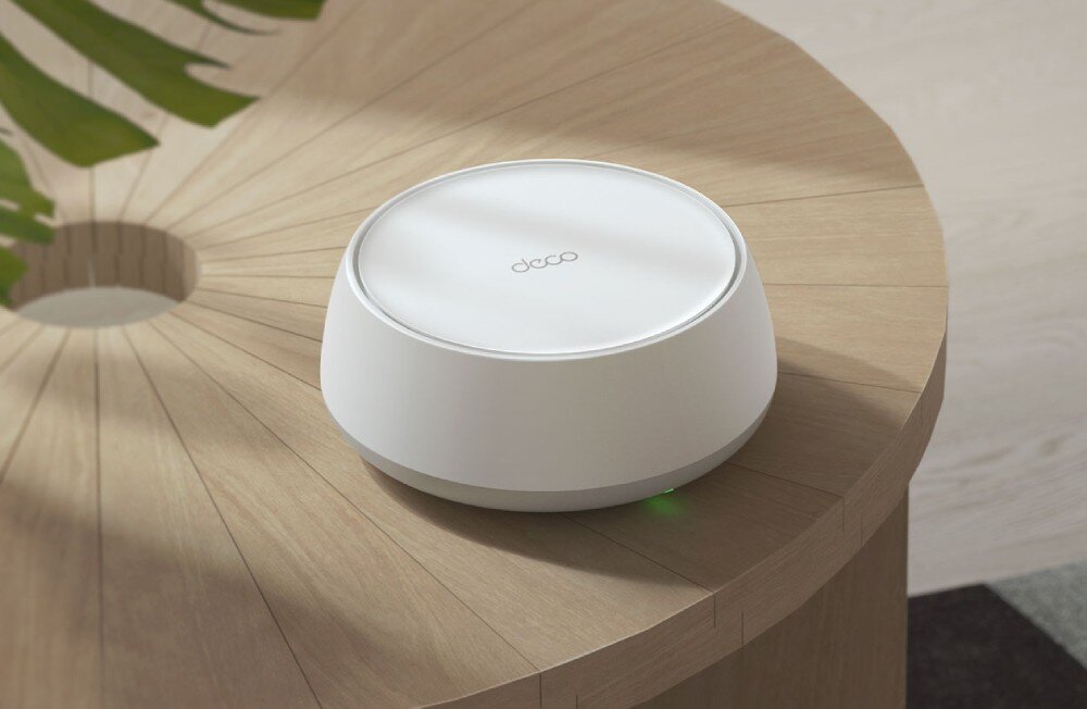 Router TP-LINK Deco BE22 Okrągłe urządzenie sieciowe z napisem 'deco' na górnej obudowie stojące na drewnianym stole obok zielonego liścia, standard, działanie, pasma