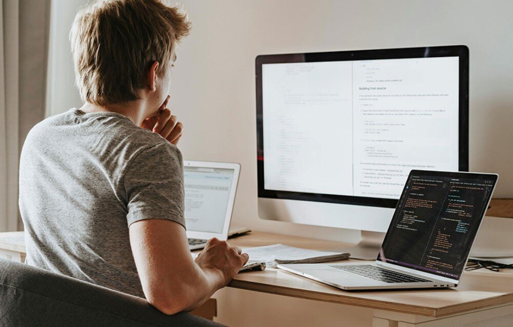Router TP-LINK Deco BE22 Mężczyzna siedzący tyłem przy biurku z trzema ekranami wyświetlającymi linie kodu programistycznego i tekst, VPN, protokoły, szyfrowanie