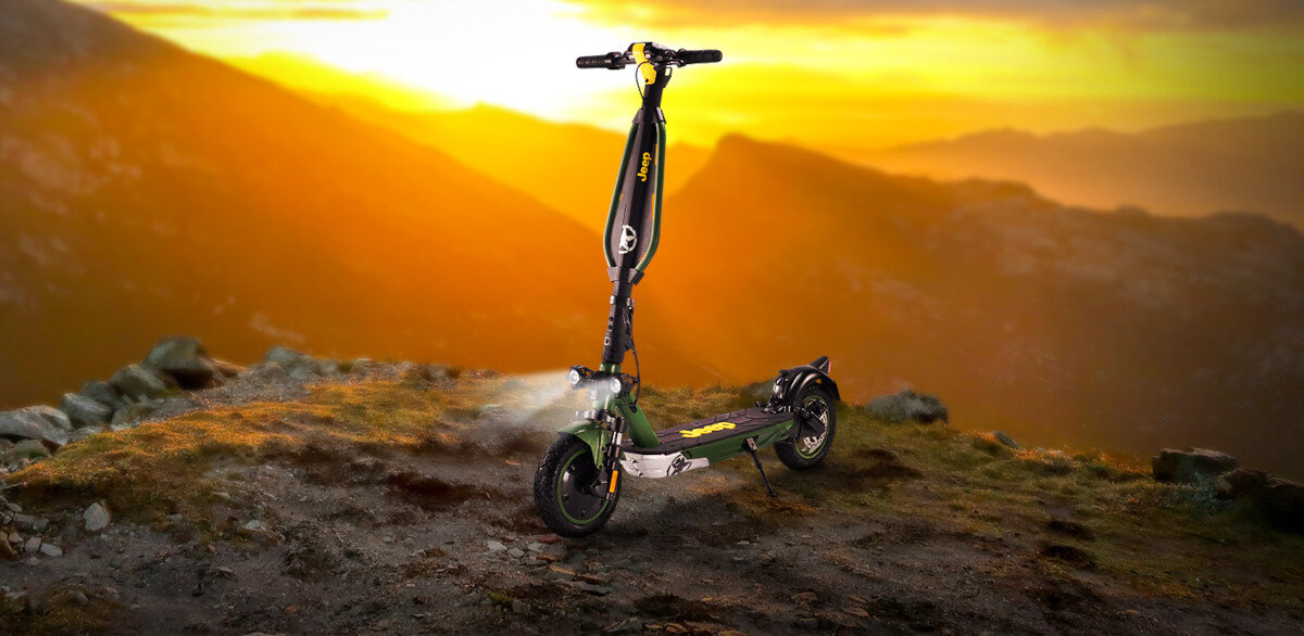 Hulajnoga elektryczna w kolorze czarno-zielonym stoi na skalistym terenie oświetlonym zachodzącym słońcem. Na kierownicy widoczne jest logo \'Jeep\'.