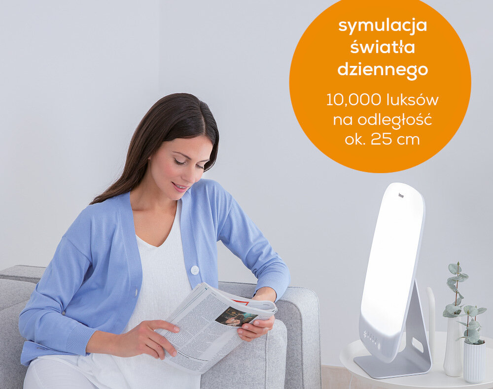 Kobieta siedzi na kanapie, trzymając magazyn, obok niej znajduje się lampa emitująca jasne światło. Na pomarańczowym tle widnieje tekst: \'symulacja światła dziennego 10,000 luksów na odległość ok. 25 cm\'.