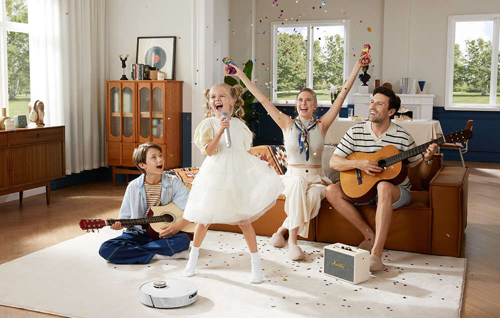 W salonie na dywanie znajduje się biały robot sprzątający. W tle widać rodzinę bawiącą się muzyką i konfetti.