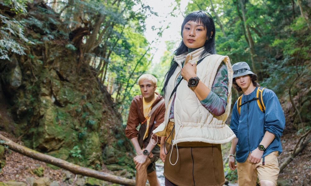 Trzy osoby stoją w lesie, każda z nich ma na ręce smartwatch. W tle widać drzewa i kamienie.