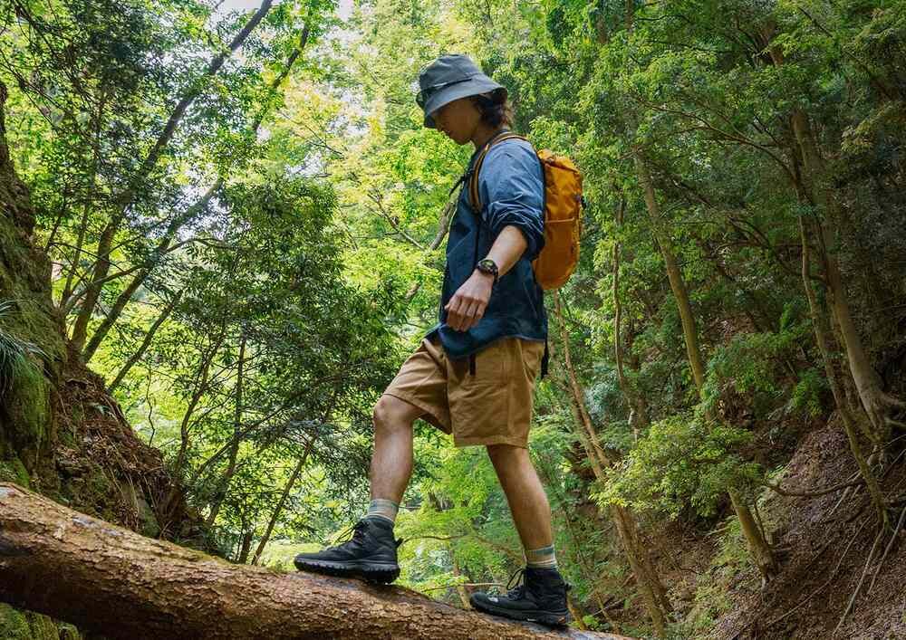 Osoba w kapeluszu i plecaku przechodzi po pniu drzewa w lesie. Na nadgarstku widoczny jest smartwatch.