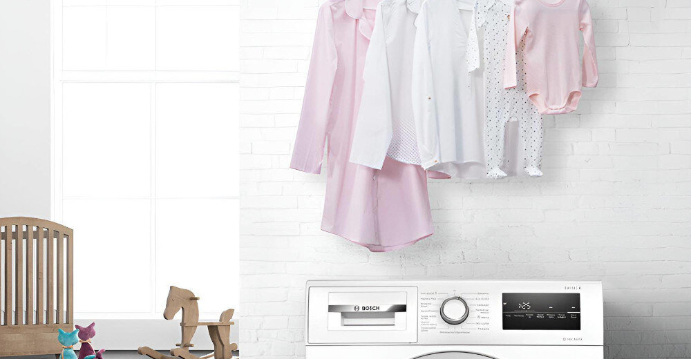Pralka z widocznym panelem sterowania i logo BOSCH, nad nią wiszą różowe i białe ubrania. W tle znajduje się biała ściana z cegieł oraz fragment drewnianego krzesła i zabawki.