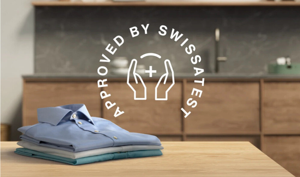 Na drewnianym blacie leży stos złożonych koszul, a w tle widać kuchenne szafki. Na obrazie znajduje się napis \'APPROVED BY SWISSATEST\' z symbolem dłoni i plusa.