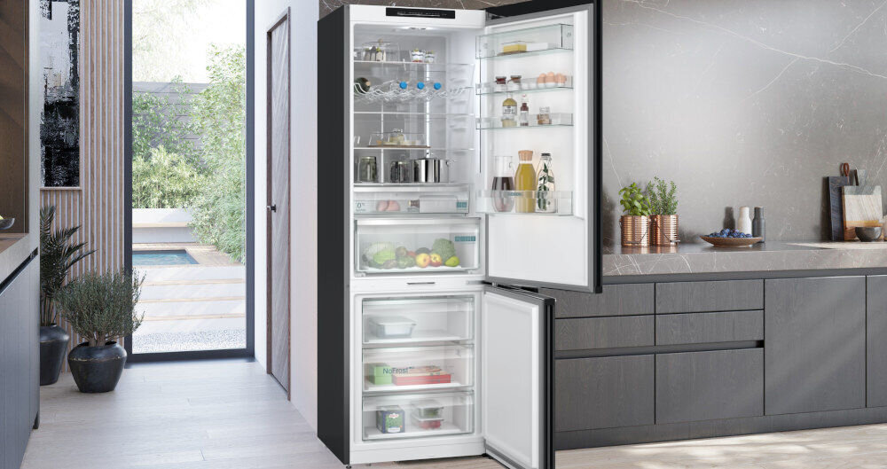 Wnętrze nowoczesnej kuchni z otwartą lodówką w zabudowie, w której widać różne produkty spożywcze na półkach. Obok lodówki znajduje się blat z dekoracjami i widok na ogród przez duże okno.