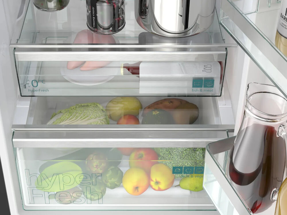 Wnętrze lodówki z widocznymi szklanymi półkami i szufladami oznaczonymi jako \'hyperFresh\'. W szufladach znajdują się różne owoce i warzywa, a na półkach widoczne są pojemniki i opakowania z jedzeniem.