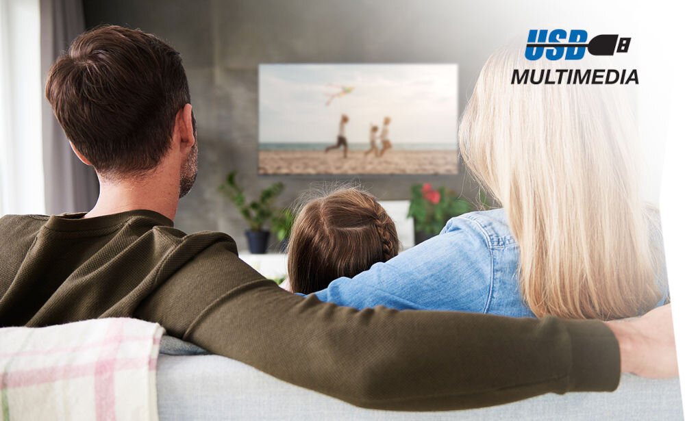 Trzy osoby siedzą na kanapie i oglądają telewizor, na którym wyświetlany jest obraz plaży z bawiącymi się dziećmi. W prawym górnym rogu widoczny jest napis \'USB MULTIMEDIA\'.