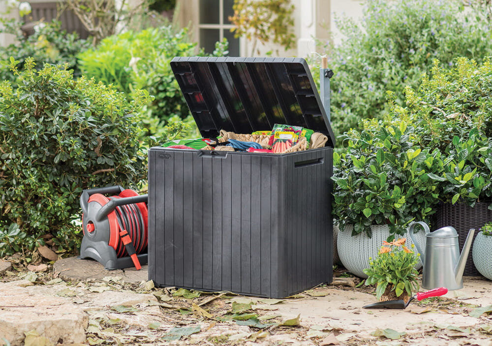 Otwarta skrzynia ogrodowa w kolorze antracytowym, wypełniona różnymi przedmiotami, stoi na tle zielonych krzewów. Obok znajduje się zwinięty wąż ogrodowy, konewka i mała roślina w doniczce.