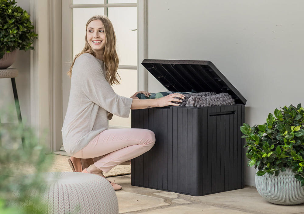 Osoba siedzi obok otwartej, antracytowej skrzyni ogrodowej, wkładając do niej złożony koc. W tle widoczne są rośliny doniczkowe i drzwi z szybami.