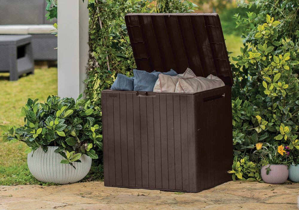 Otwarta skrzynia ogrodowa w kolorze antracytowym, wypełniona poduszkami w różnych kolorach, stoi na tarasie otoczonym roślinami. W tle widoczne są zielone krzewy i fragment mebli ogrodowych.