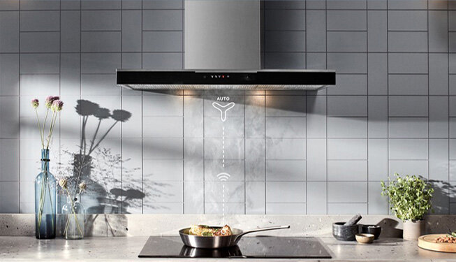 Na obrazie widoczny jest czarny okap kuchenny zamontowany nad płytą grzewczą, na której stoi patelnia z jedzeniem. Na ścianie za okapem znajduje się napis \'AUTO\' oraz symbole związane z funkcjami urządzenia.