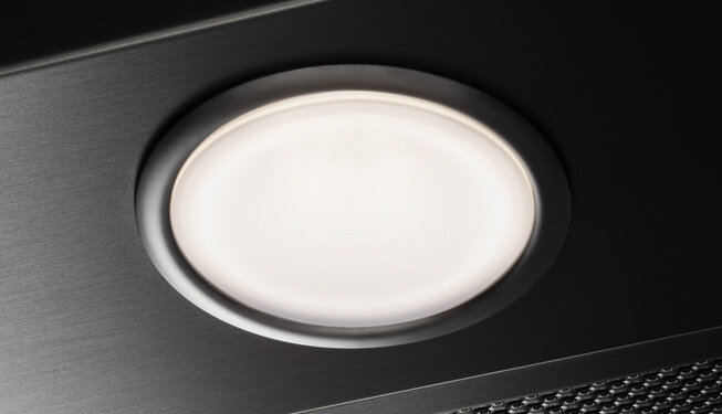 Zbliżenie na okrągłe, wbudowane światło w czarnym okapie kuchennym. Obok widoczna jest część metalowej kratki wentylacyjnej.