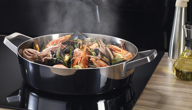 Na płycie grzewczej stoi metalowy garnek z uchwytami, w którym gotują się owoce morza. Obok znajduje się butelka z oliwą i metalowy młynek do przypraw.