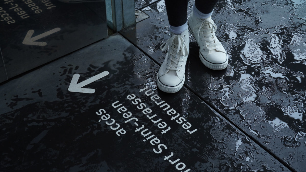 Osoba w białych trampkach stoi na mokrej powierzchni z napisami: \'accès port Saint-Jean terrasse restaurant\' oraz strzałkami wskazującymi kierunek.