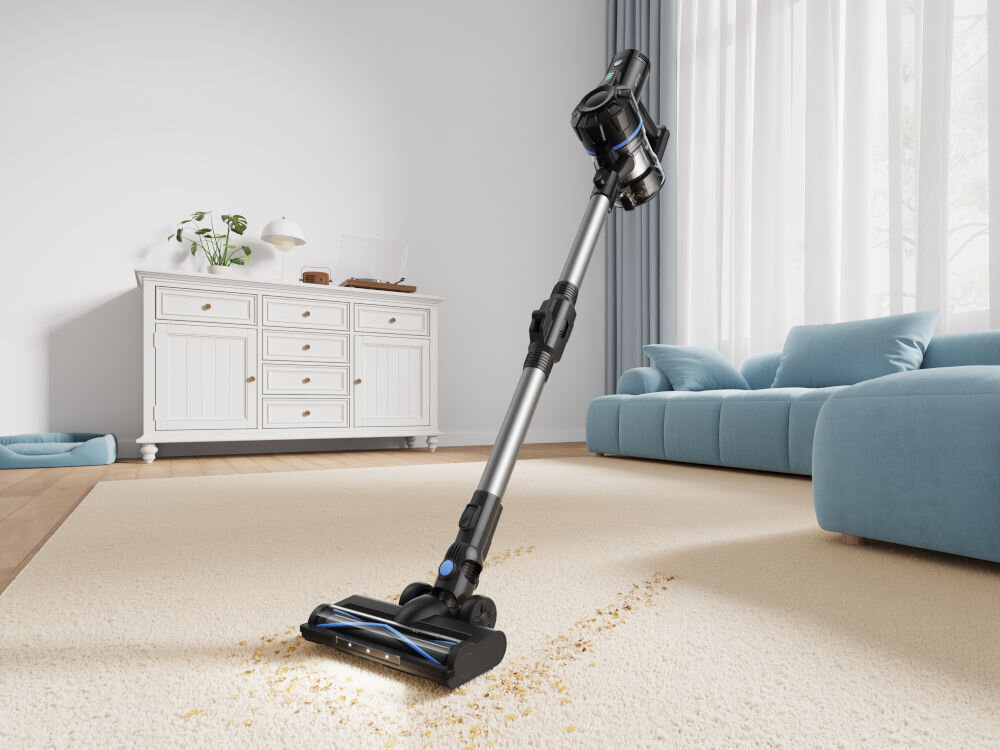Odkurzacz pionowy z metalową rurą i czarną głowicą czyszczącą stoi na jasnym dywanie w salonie. W tle znajduje się biała komoda z rośliną i niebieska sofa przy oknie z zasłonami.