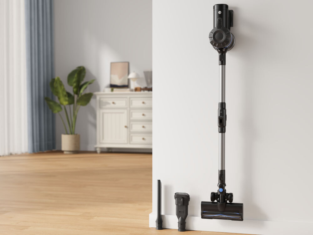 Na zdjęciu widać odkurzacz pionowy zamontowany na ścianie oraz dwie dodatkowe końcówki leżące na podłodze. W tle znajduje się komoda i roślina w doniczce.