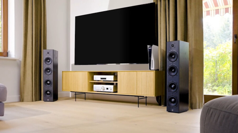 W salonie znajduje się duży telewizor na drewnianej szafce, obok której stoją dwie czarne kolumny głośnikowe. Pomieszczenie jest jasne, z dużymi oknami i zasłonami.