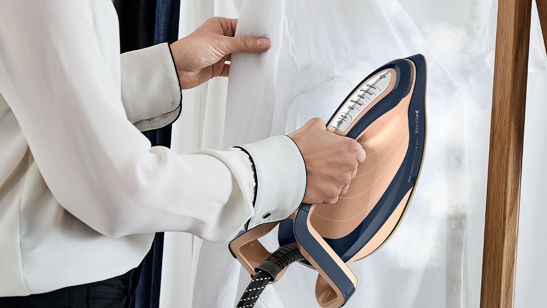 Osoba prasuje białą tkaninę przy użyciu generatora pary w kolorze granatowo-miedzianym. Urządzenie emituje parę, a na jego powierzchni widoczne są przyciski i otwory parowe.