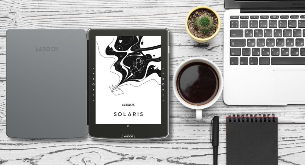Na drewnianym stole znajduje się otwarte etui z urządzeniem INKBOOK, na ekranie widoczny jest napis \'SOLARIS\'. Obok leży laptop, filiżanka z kawą, notatnik, długopis i mała doniczka z kaktusem.