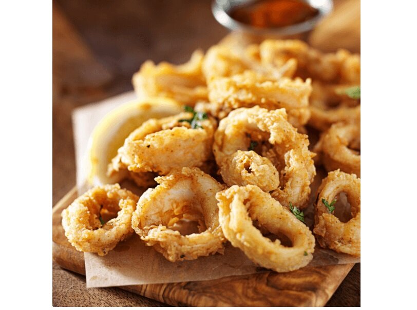 Na obrazie widoczne są smażone kalmary ułożone na papierze do pieczenia. W tle znajduje się niewyraźny sos w małej miseczce.