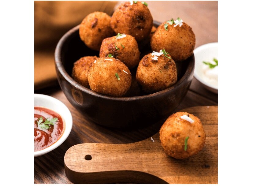 Na drewnianej desce znajduje się miska z kilkoma złocistymi kulkami jedzenia, obok miski widoczny jest sos w małej miseczce. Kulki są posypane drobnymi ziołami i startym serem.