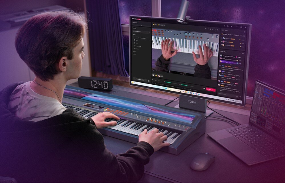 Osoba gra na elektronicznym pianinie, patrząc na monitor z logo \'YOGA\', na którym wyświetlany jest obraz rąk grających na klawiaturze. Obok znajduje się laptop i myszka, a na pianinie stoi zegar pokazujący godzinę 12:40.