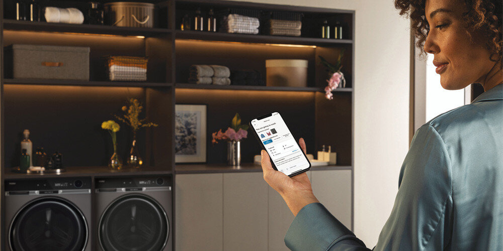 PRALKA HAIER HW100BD14397PGUS X11 lifestyle wizualizacja kobieta z telefonem w dłoni aktywna aplikacja wyświetlacz dwa urządzenia w tle garderoba półki ubrania odzież Zdalne sterowanie z aplikacją hOn 60 dodatkowych programów informacje powiadomienia kontrola smartfon tablet telefon zdalne Zarządzanie