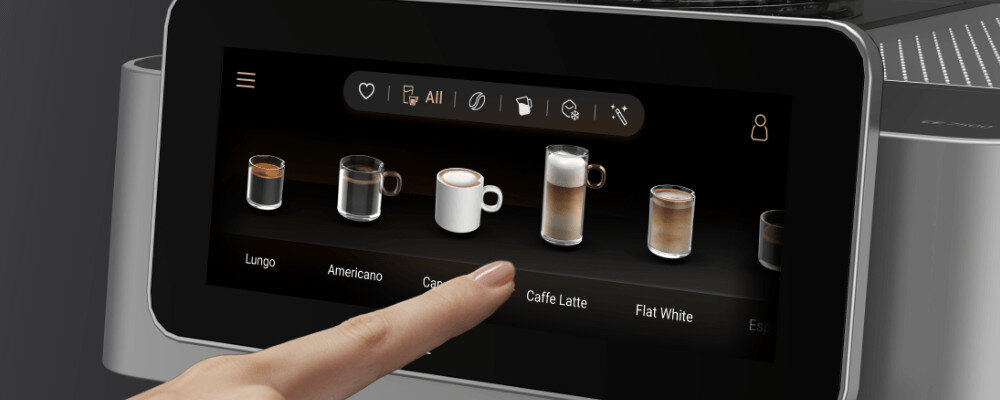 Ekran dotykowy ekspresu do kawy pokazuje różne opcje napojów, takie jak Lungo, Americano, Cappuccino, Caffe Latte i Flat White. Palec wskazuje na jedną z opcji na ekranie.
