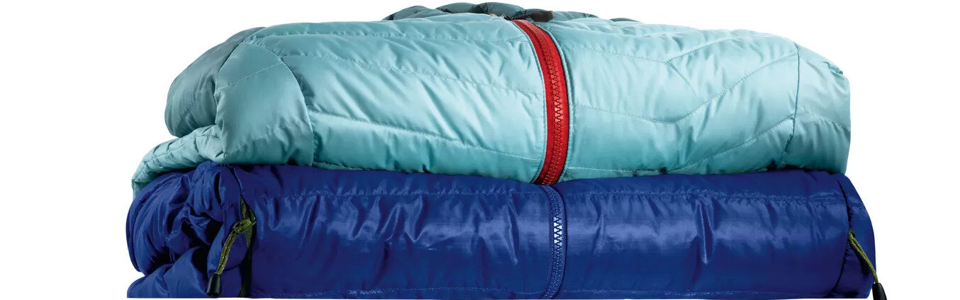 Na obrazie widoczne są dwa złożone śpiwory, jeden w kolorze jasnoniebieskim z czerwonym zamkiem, a drugi w kolorze ciemnoniebieskim z niebieskim zamkiem.