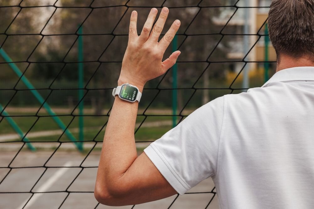Osoba w białej koszulce stoi przy siatce, na nadgarstku ma zielony smartwatch z wyświetlaczem pokazującym dane. W tle widać rozmyty krajobraz z elementami zieleni i budynków.