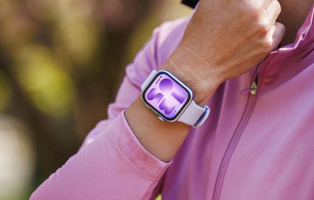 Osoba w różowej bluzie nosi smartwatch z fioletowym wyświetlaczem na nadgarstku.