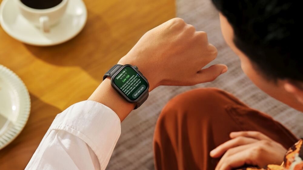 Osoba siedzi przy stole, patrząc na smartwatch na nadgarstku. Na ekranie zegarka widoczny jest tekst w języku angielskim.