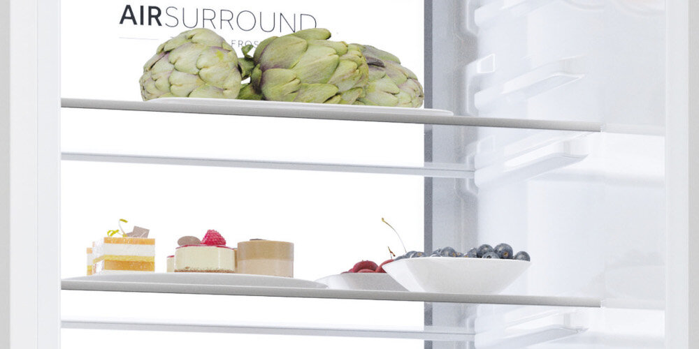 Wnętrze lodówki z trzema półkami, na których znajdują się karczochy, ciasta i miska z jagodami. Widoczny jest napis \'AIR SURROUND\'.