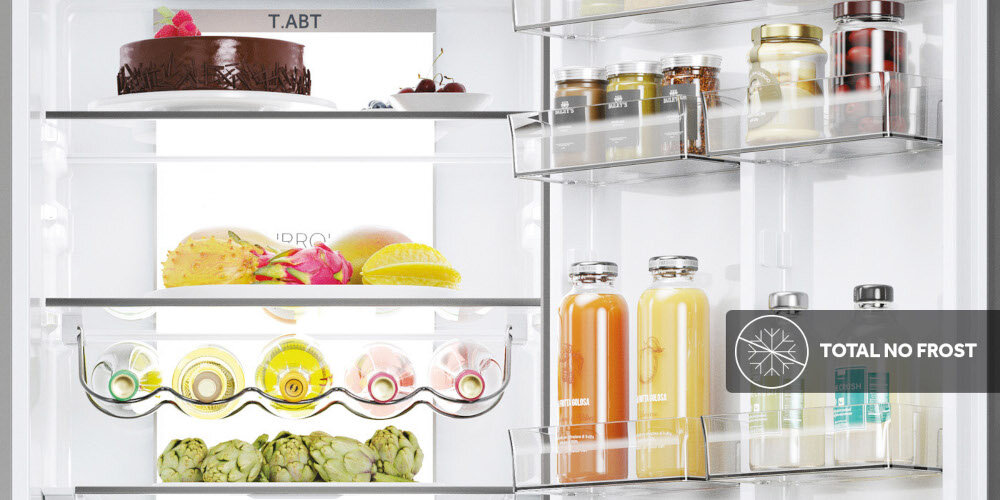 Wnętrze lodówki z widocznymi półkami, na których znajdują się różne produkty spożywcze, takie jak ciasto, owoce, soki i słoiki. Na drzwiach lodówki widoczny jest napis \'TOTAL NO FROST\'.