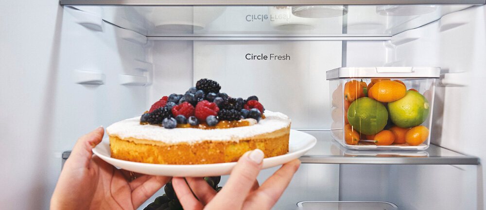 Wnętrze lodówki z widocznym ciastem z owocami trzymanym w dłoniach oraz pojemnikiem z owocami cytrusowymi na półce. Na tylnej ściance widoczny jest napis \'Circle Fresh\'.