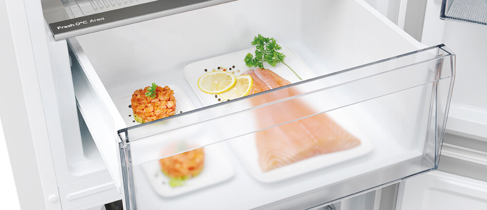 Otwarta szuflada lodówki z przezroczystym frontem, w której znajdują się dwa talerze z jedzeniem: plasterki łososia z cytryną i natką pietruszki oraz porcja siekanego mięsa. Na górnej krawędzi szuflady widoczny napis \'Fresh OFC Area\'.