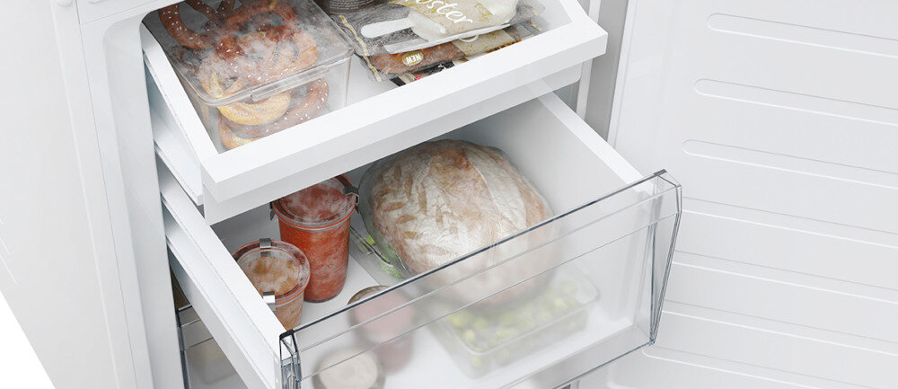 Otwarta lodówka z widocznymi dwoma szufladami, w których znajdują się zamrożone produkty spożywcze, w tym pieczywo i słoiki z przetworami. W górnej szufladzie widoczne są również opakowania z mrożonymi przekąskami.