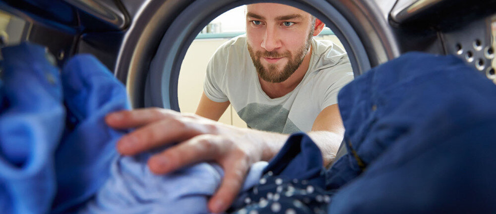 Osoba wkłada ubrania do bębna pralki. Widać różnokolorowe tkaniny i metalowe wnętrze urządzenia.